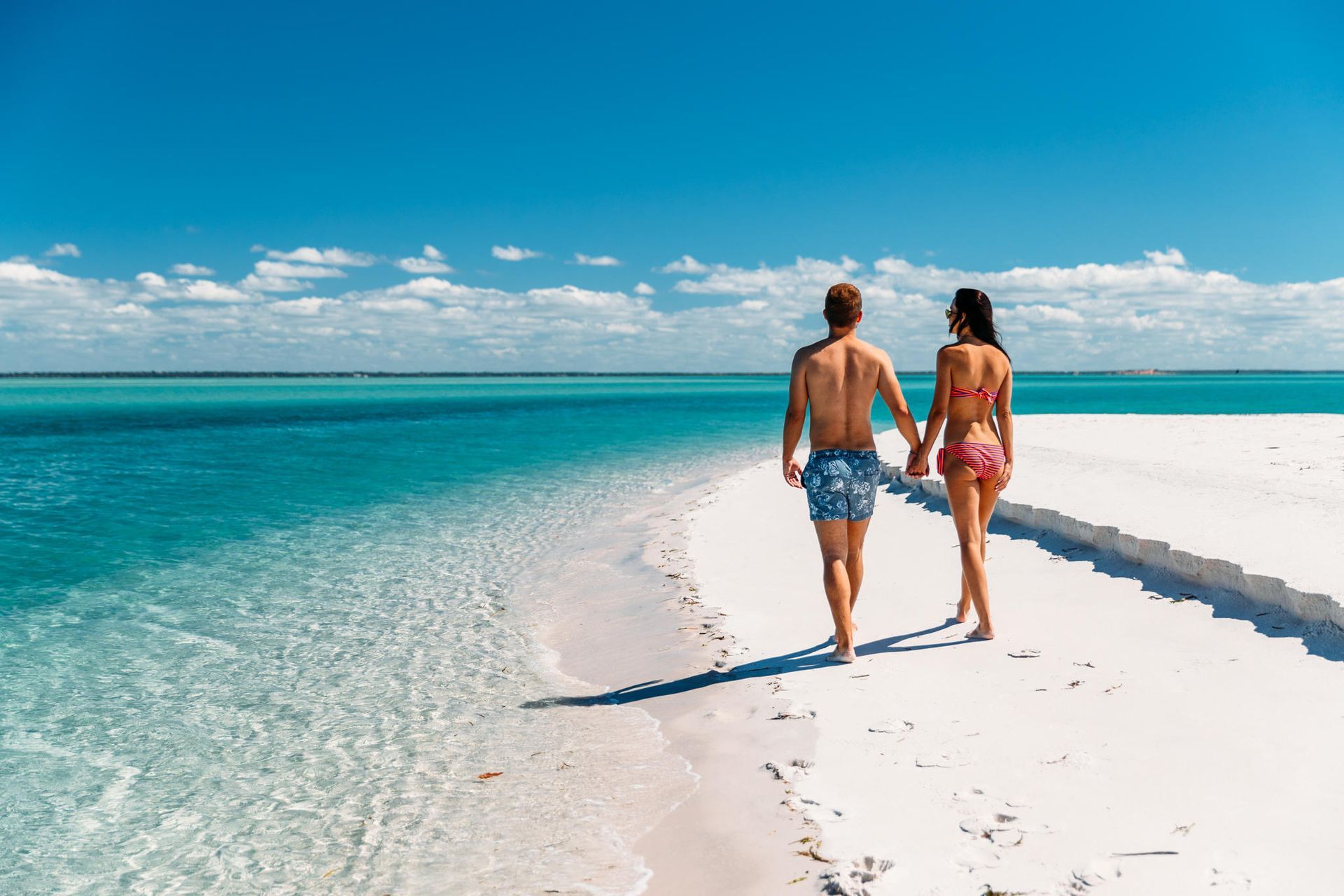 Two people walking on a white sandbar beside turquoise water under a bright blue sky