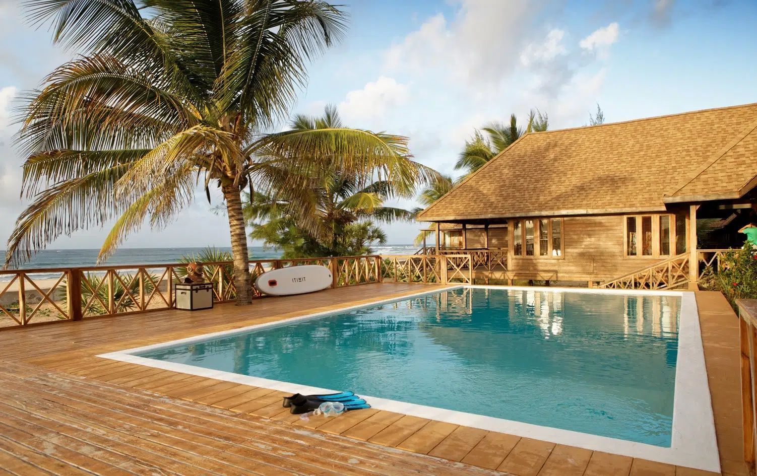 A rectangular swimming pool on a wooden deck next to a thatched-roof house, with palm trees and an ocean view nearby.