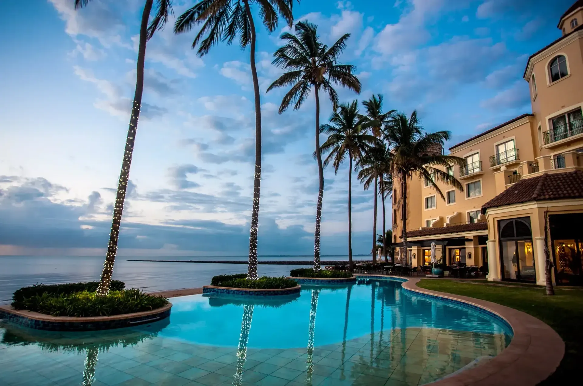 Palm-lined resort pool beside the ocean at sunset, with a beige hotel and blue sky reflecting in the water
