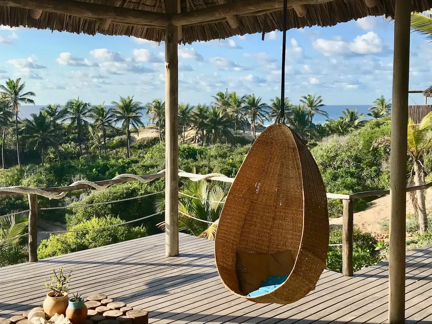 Tropical patio with woven hanging chair, thatched roof, and ocean-view palm trees beyond a garden path