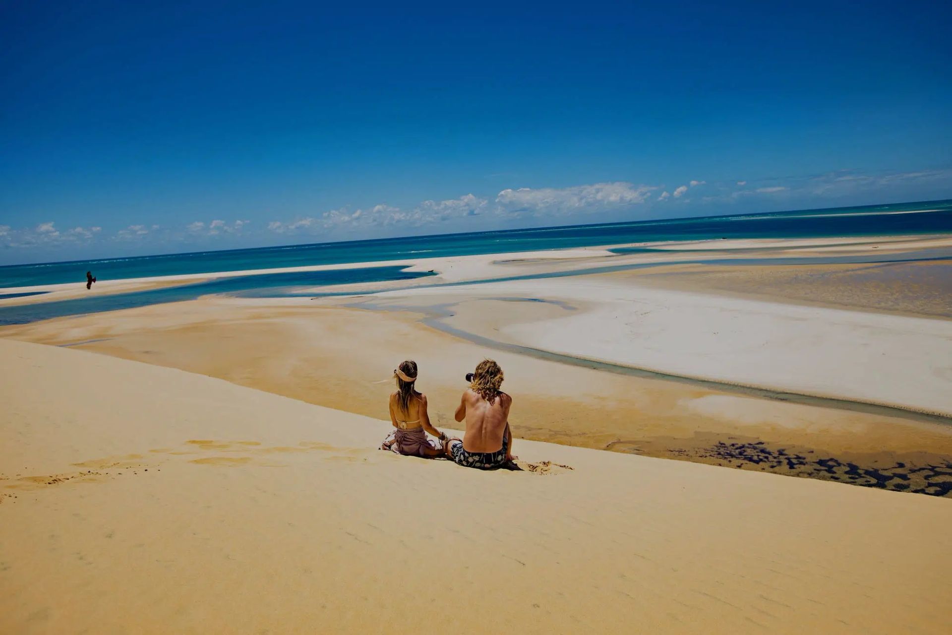 Two people sitting on a sandy beach overlooking blue ocean and tidal flats under a clear sky