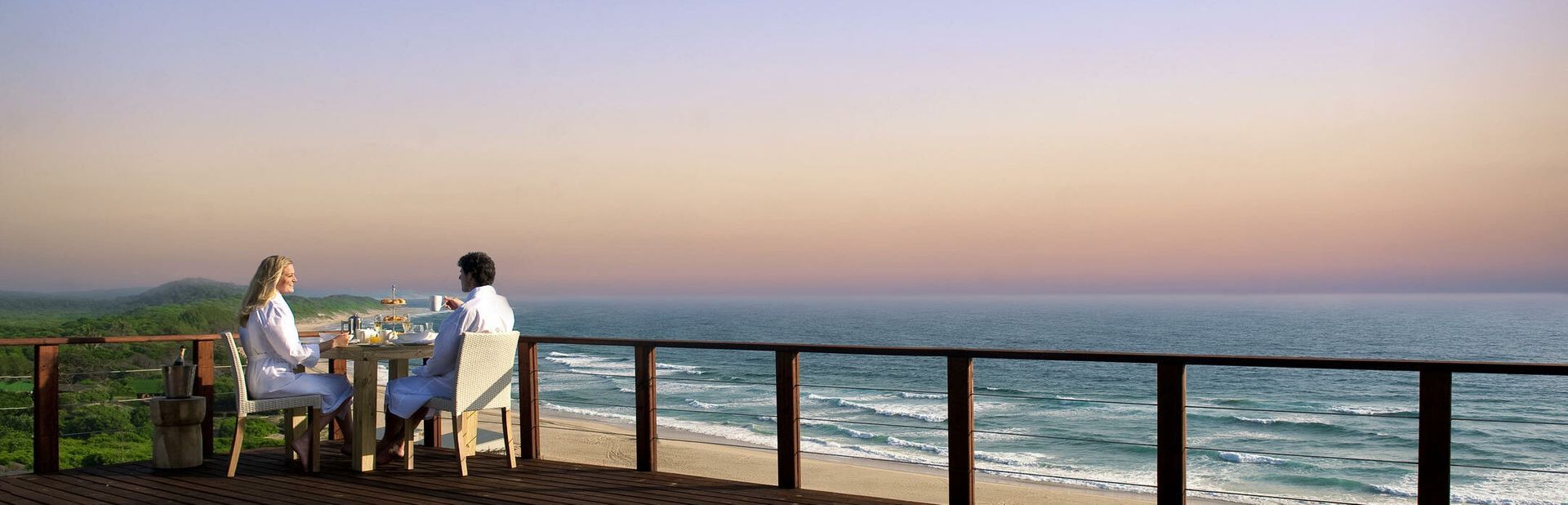 Two people sit at a seaside patio table watching the ocean at sunset.