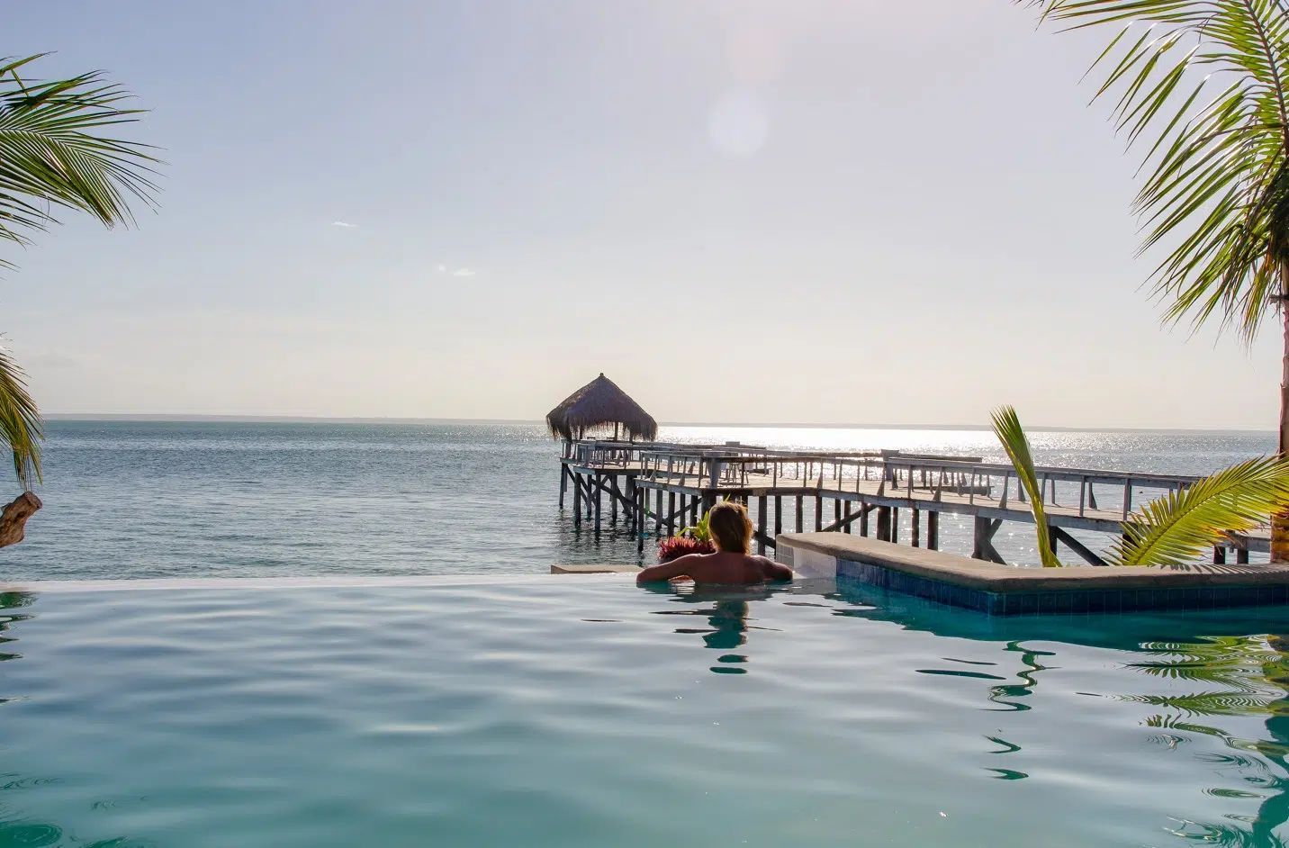 Infinity pool overlooking the ocean with a thatched pier and small boat at sunset