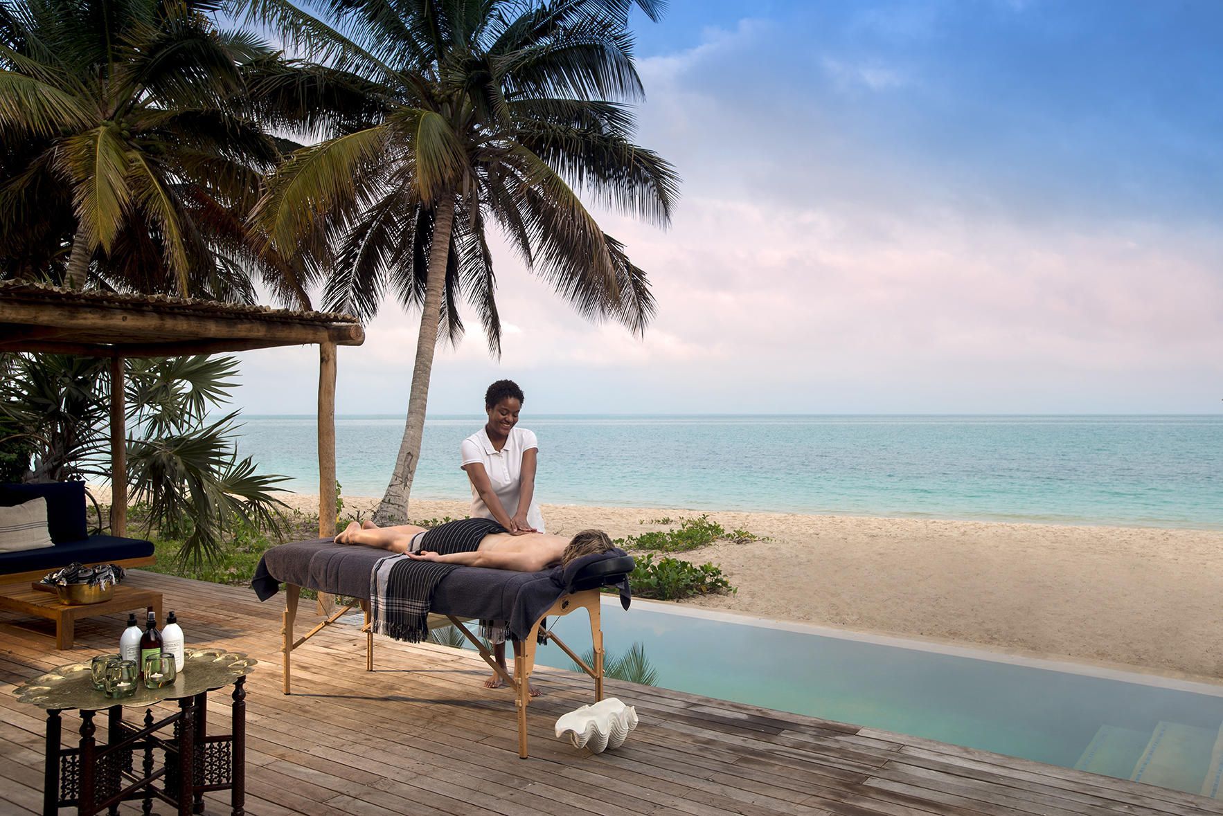 Beachside massage table under palm trees beside a pool, with ocean and sunset in the background