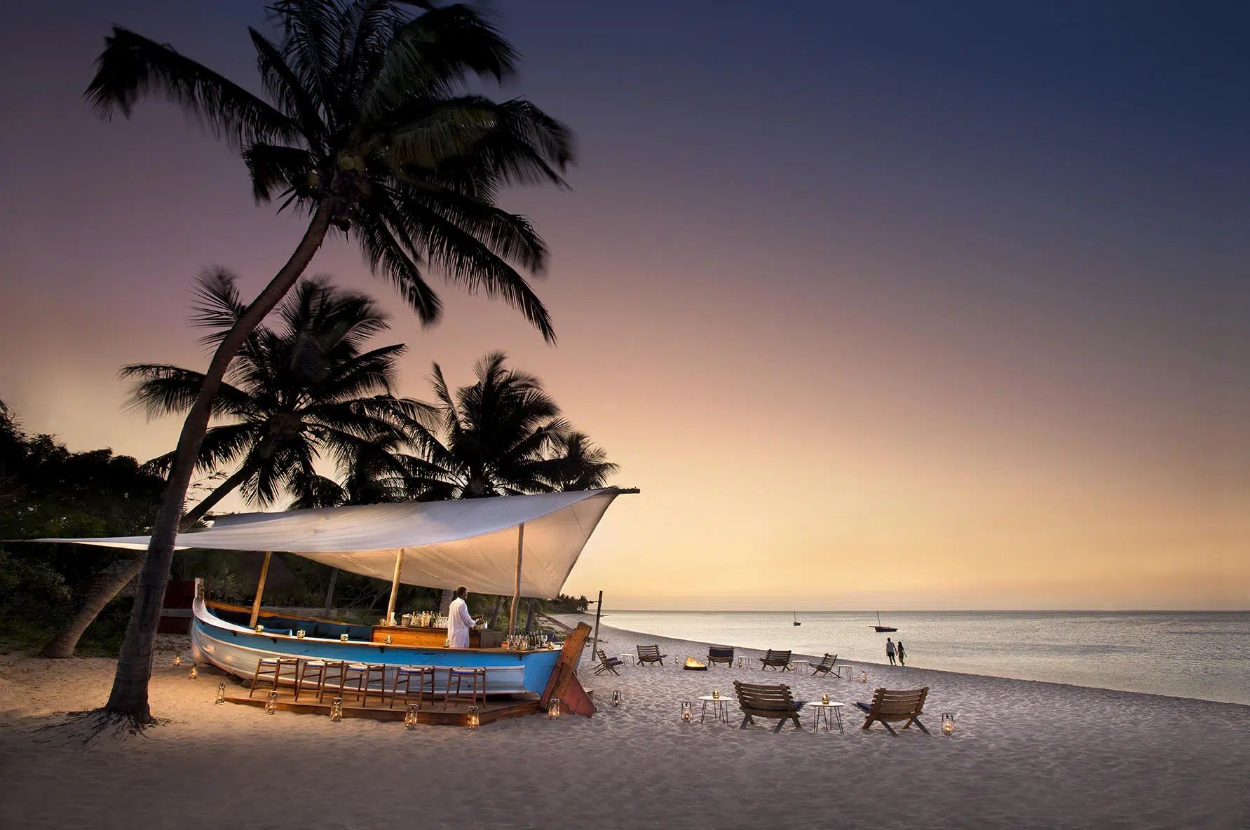 Beach at sunset with palm trees, a small boat, and lounge chairs by calm water