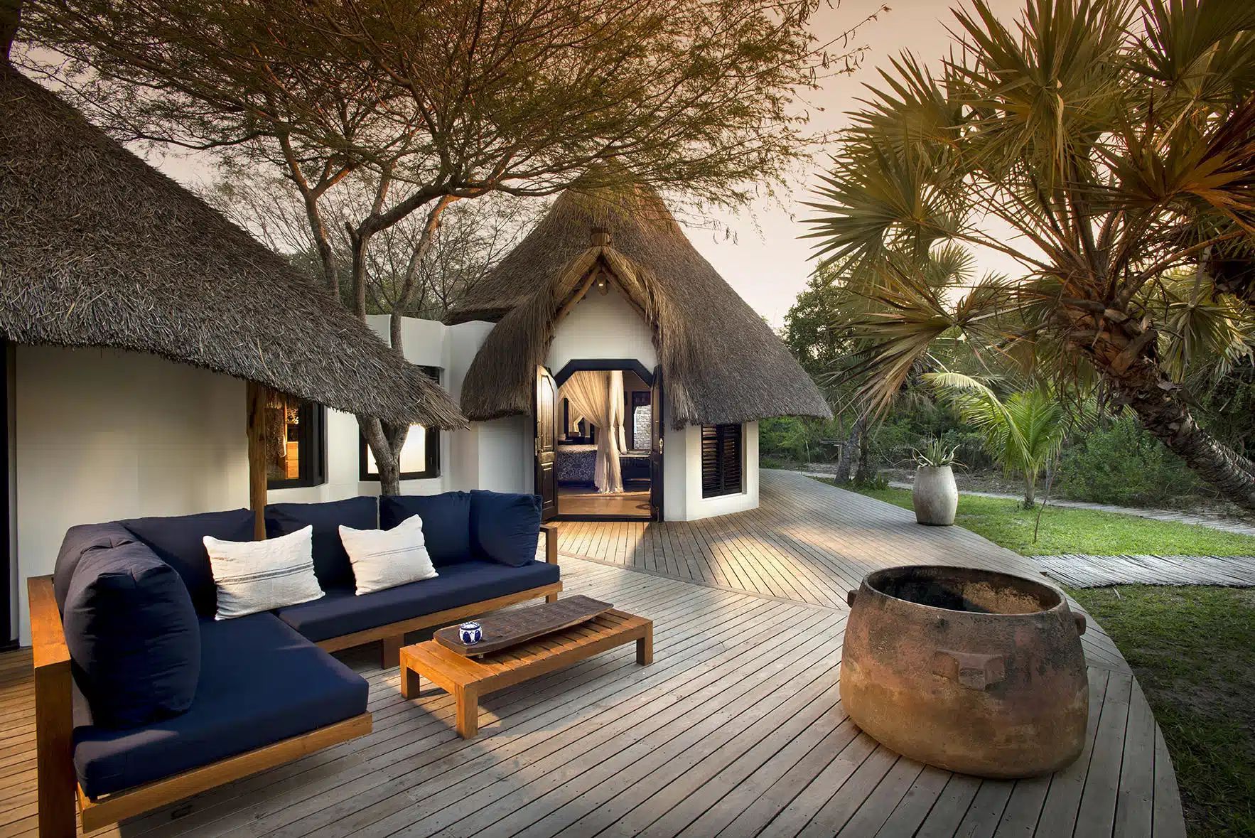 Thatched outdoor lounge with dark blue sofa, wooden deck, fire pit, and tropical trees at sunset
