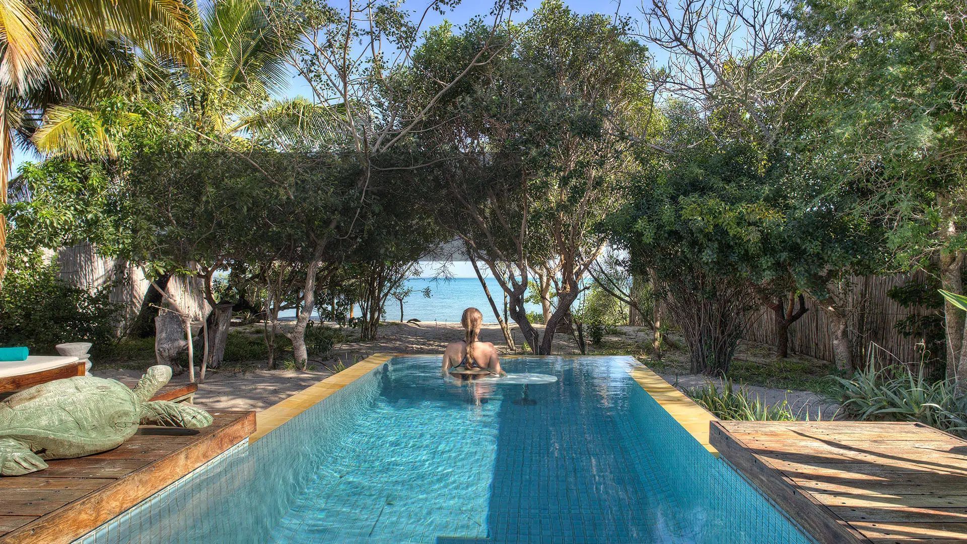 Person floating in a turquoise pool surrounded by lush trees and a sunlit deck, with ocean visible beyond.