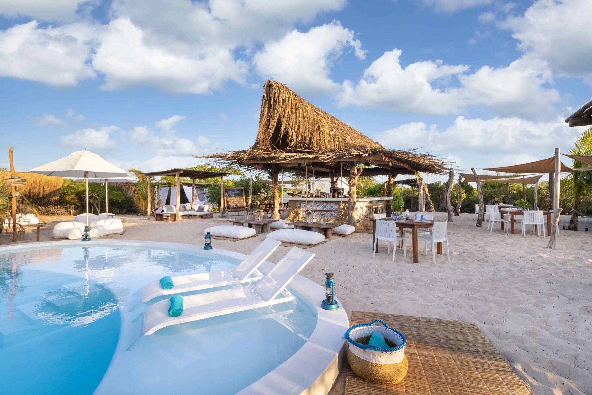 Sunlit poolside resort with white loungers, thatched shade structures, and ocean view