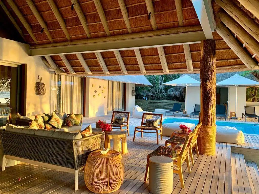 Open-air beach lounge with wicker seating, thatched roof, and a pool overlooking the ocean at sunset