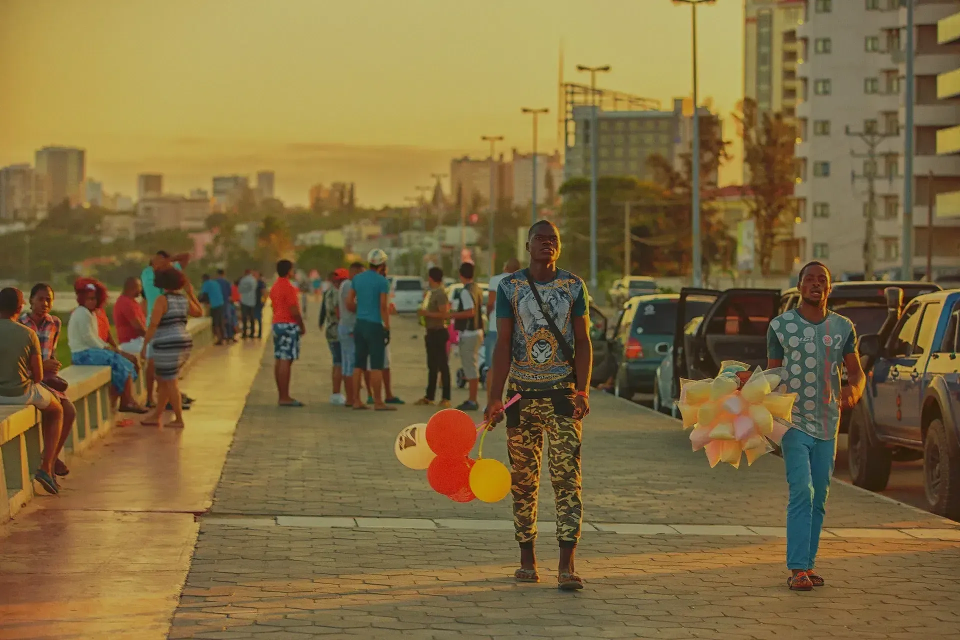People walking along a sunlit promenade, with balloons and cars parked beside the path.