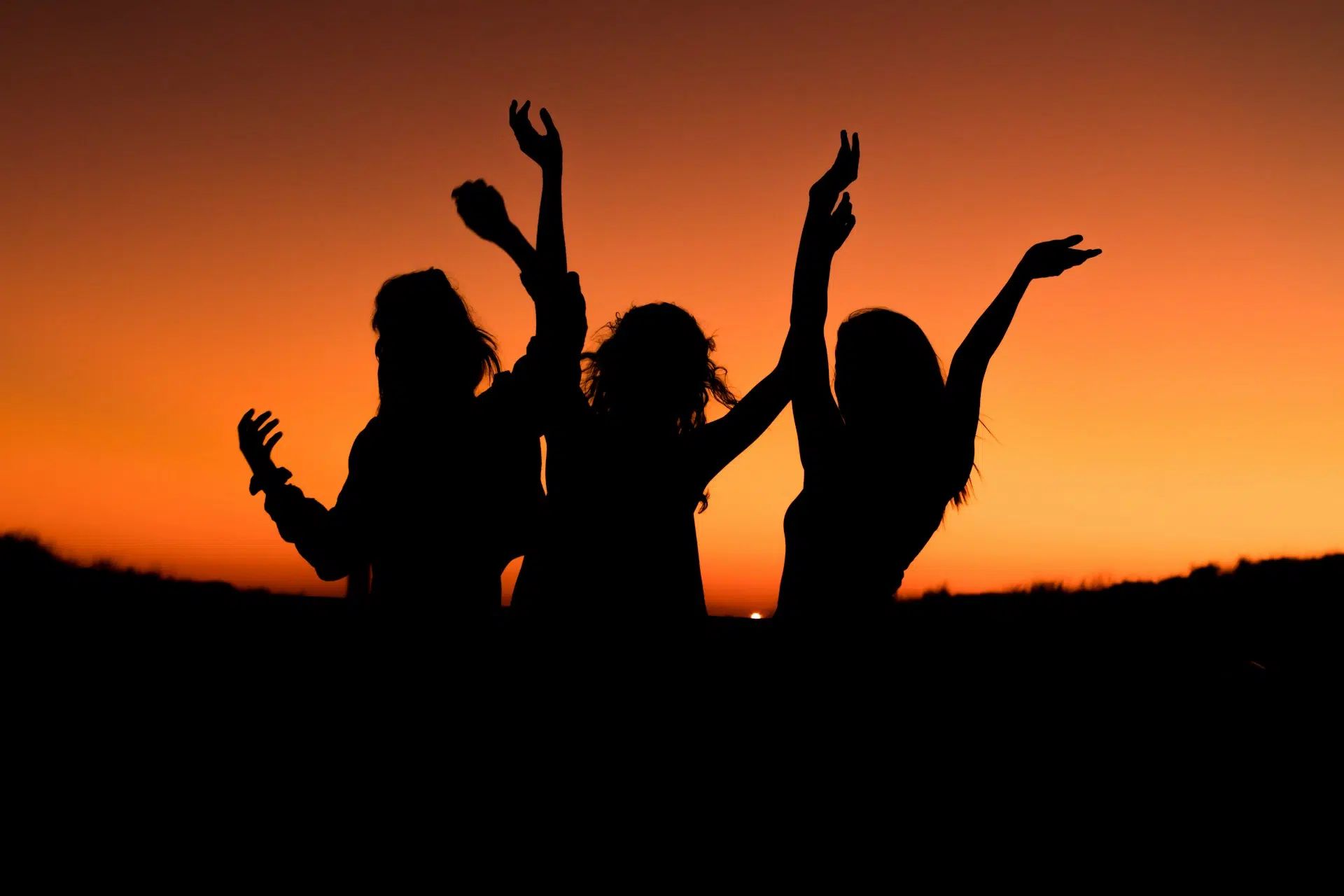 Silhouettes of four people dancing with raised arms against an orange sunset sky