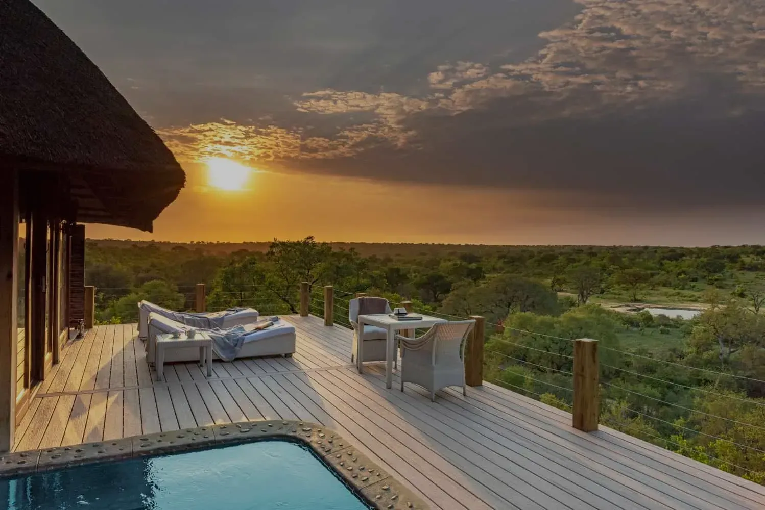 Sunset view from a deck with pool, lounge chairs, and dining table overlooking green landscape