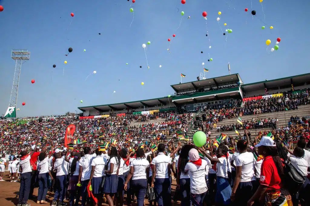A large crowd in a stadium releases many colorful balloons into a bright blue sky.
