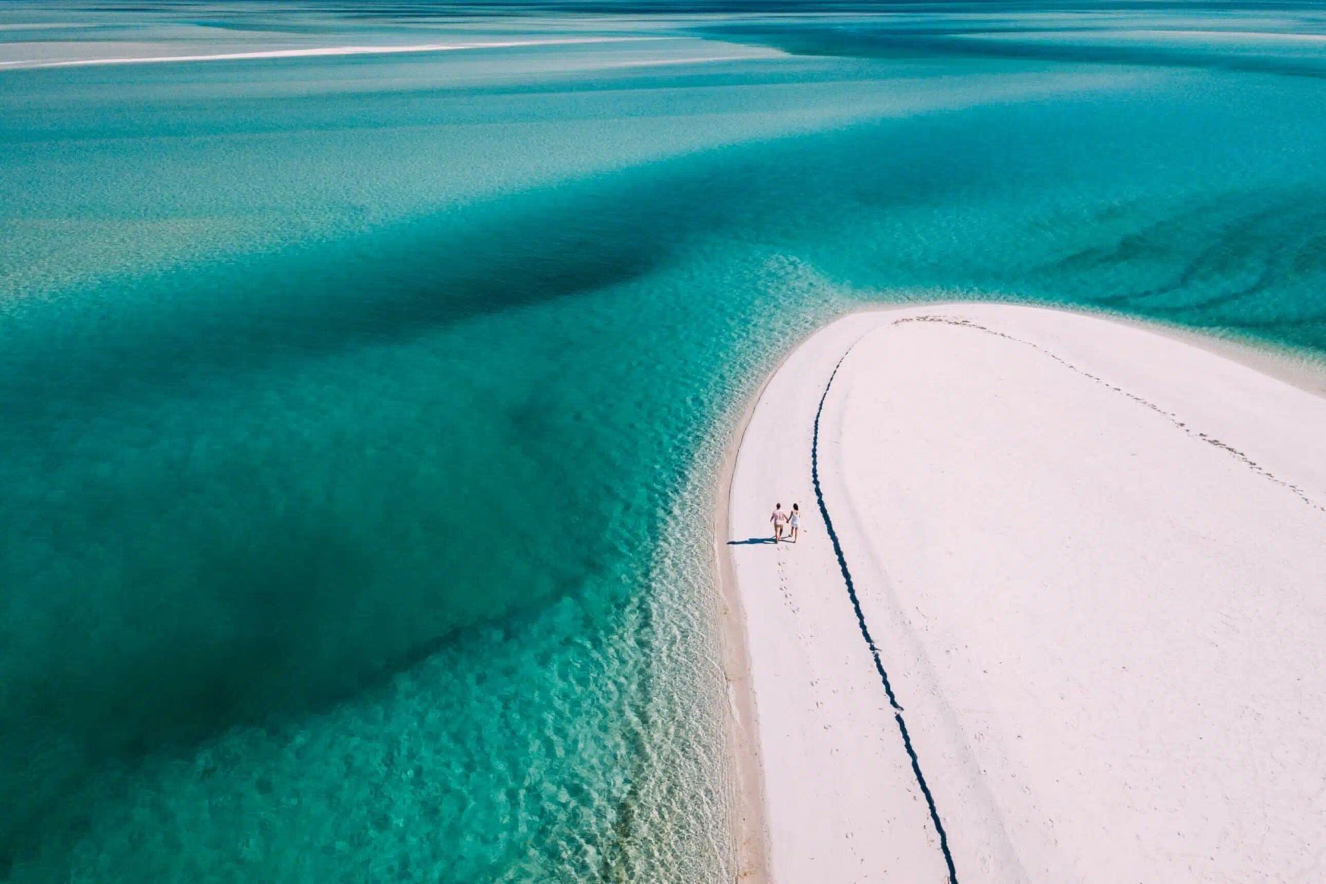 An aerial view of a person standing on a curved, white sandbar surrounded by vibrant turquoise tropical water.
