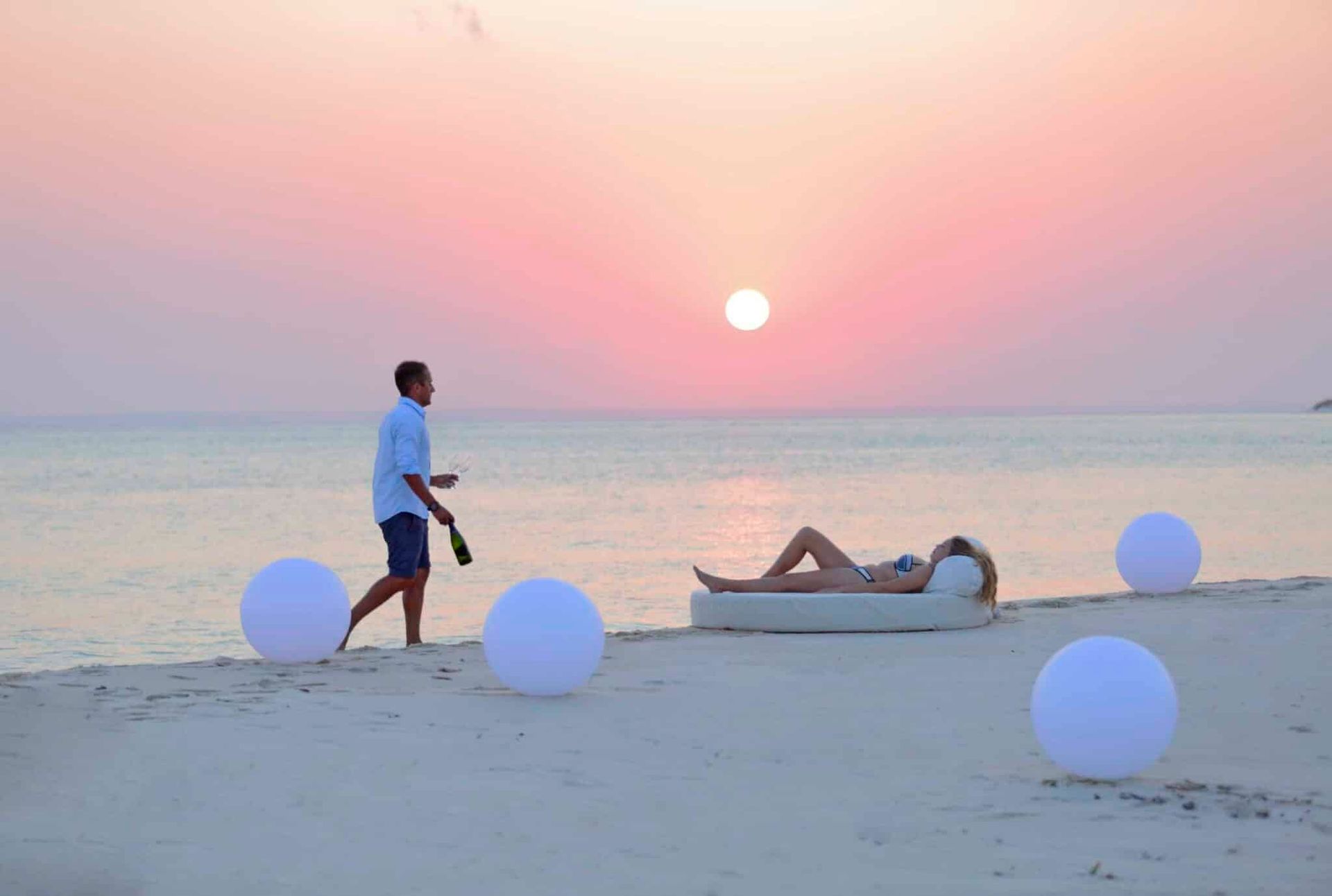 A person walks along a sandy beach at sunset, carrying a bottle toward a reclining figure on a circular lounge bed.