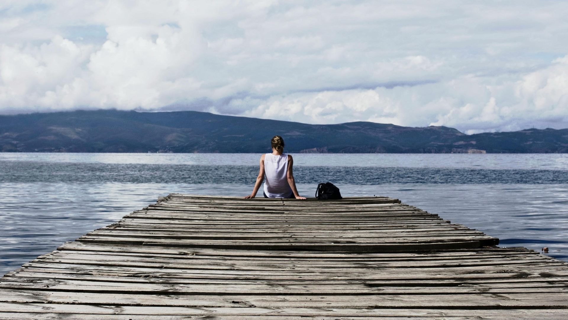 Person sitting at the end of a wooden dock, facing a calm lake and distant mountains under cloudy skies