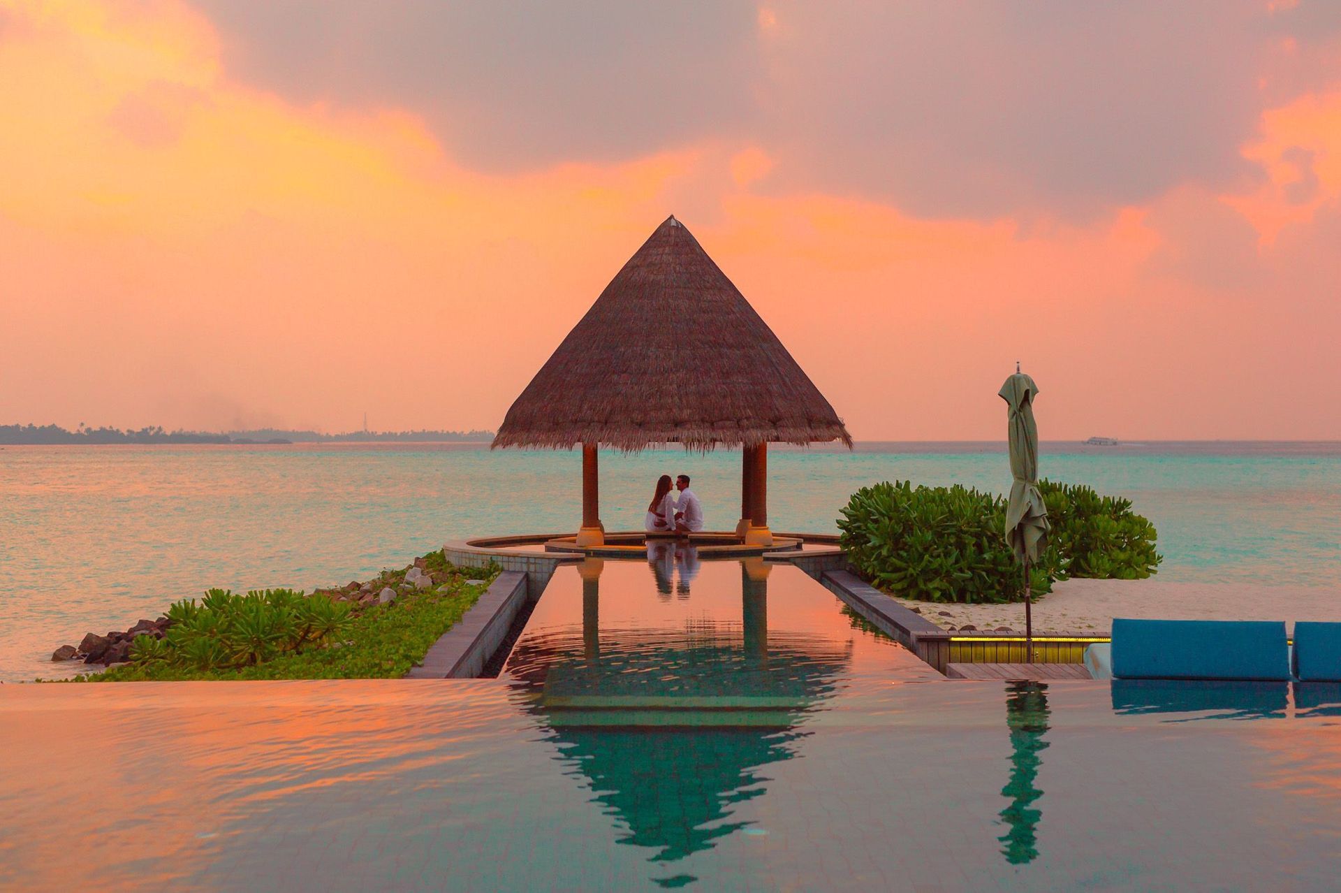 Two people sit on a dock gazebo at sunset over calm water, with an infinity pool in the foreground.