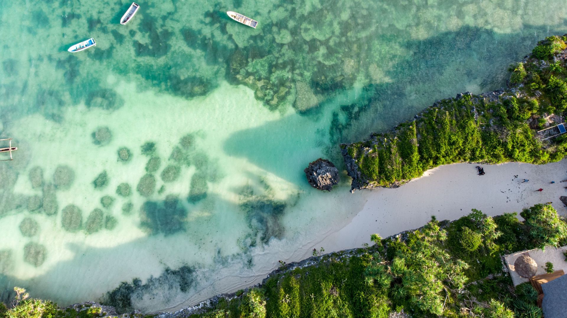 Aerial view of a tropical beach with clear turquoise water, small boats, and dense green shoreline