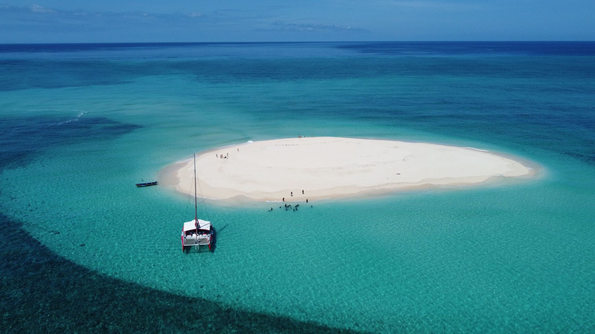 Small white sand island in turquoise ocean with a sailboat nearby