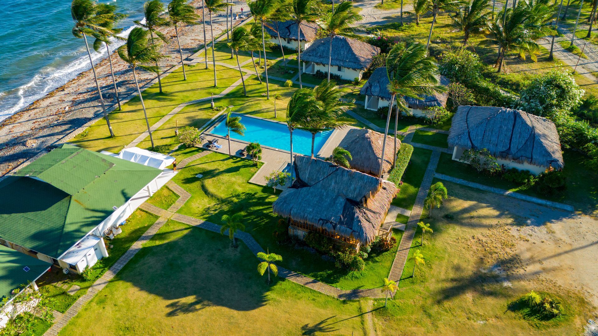 Aerial view of beachfront thatched cottages with palm trees and sandy paths near turquoise water.