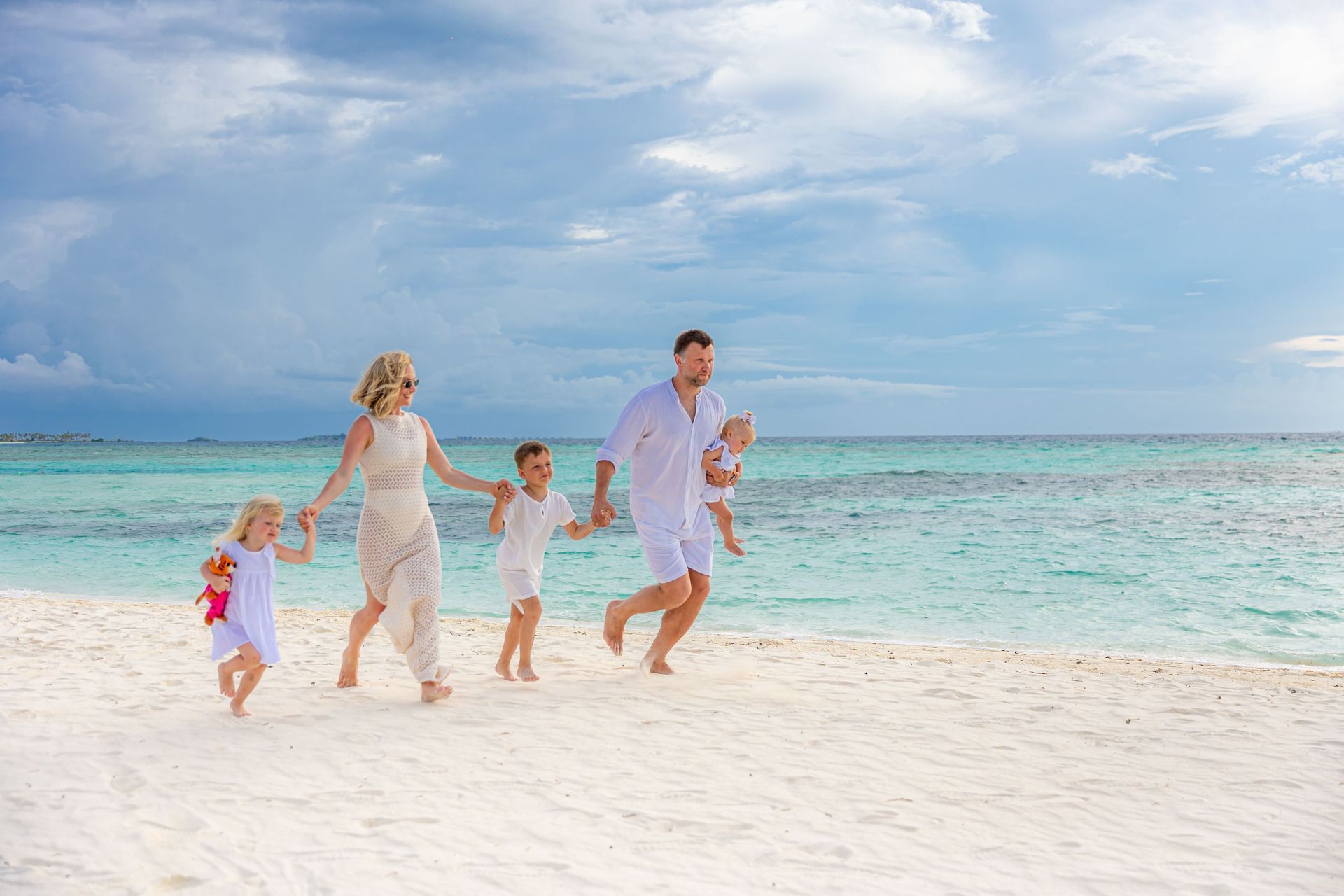 Family running on a sunny white-sand beach beside turquoise water and a cloudy sky