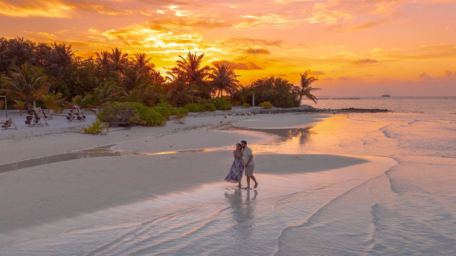 Two people walking along a tropical beach at sunset with palm trees and glowing orange sky.