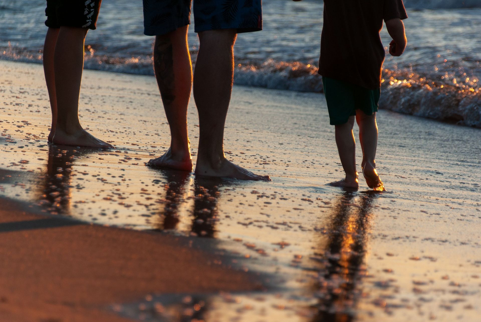 Three people standing barefoot at the shoreline as waves wash over the beach at sunset