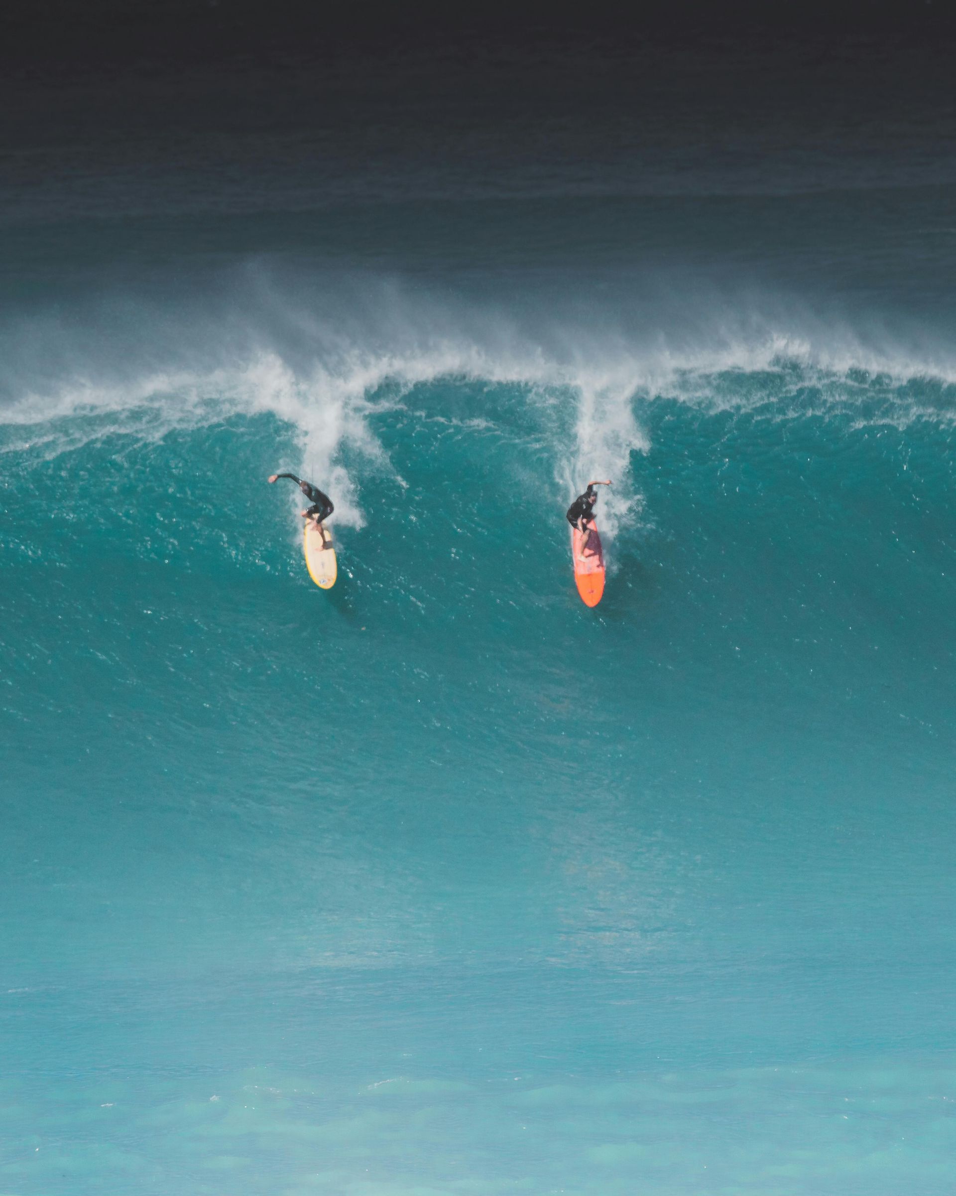 Two surfers dropping into a turquoise wave from above, with white spray and dark water behind them