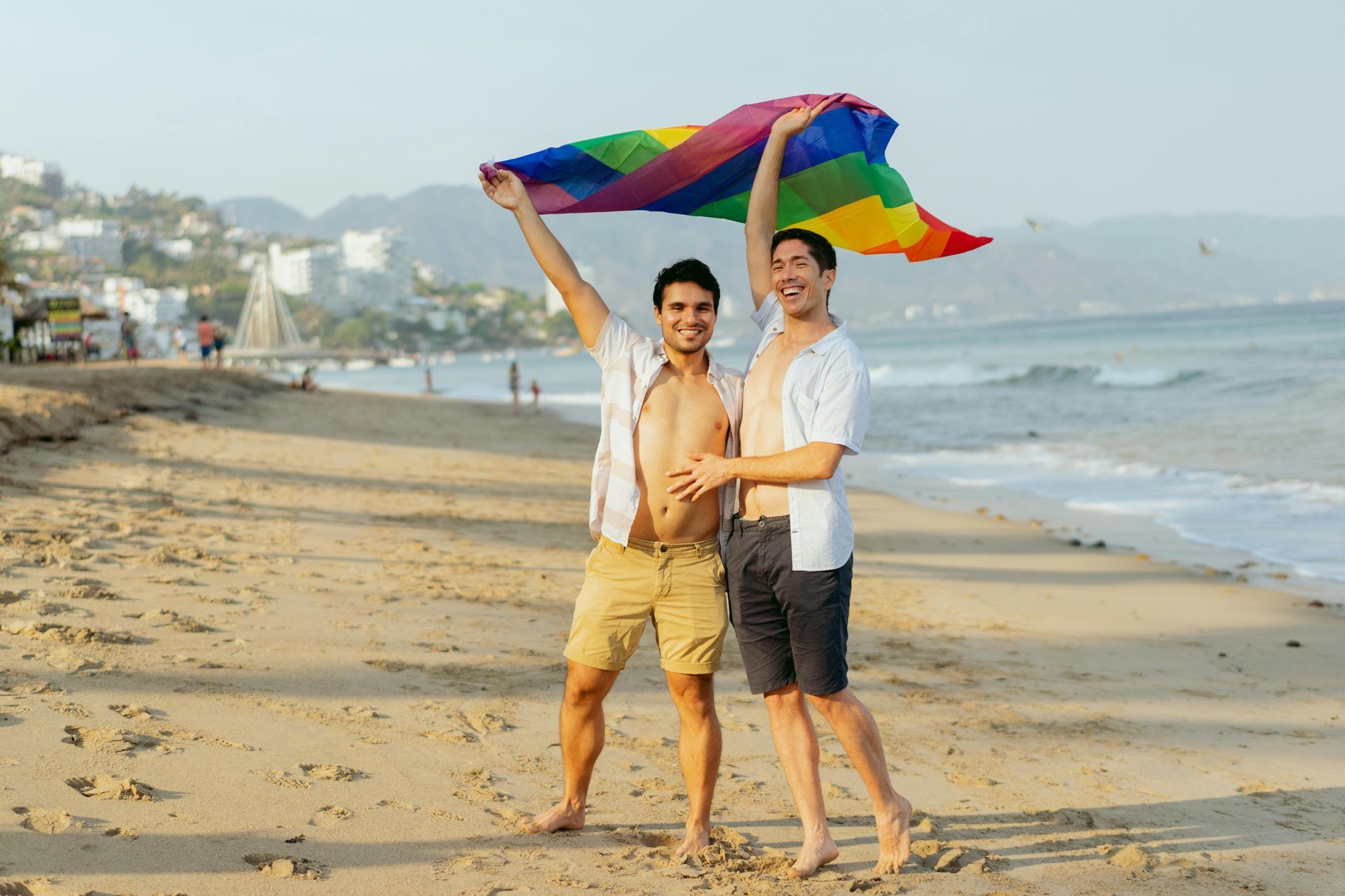 Two people holding a rainbow flag on a beach, standing by the shoreline with waves and mountains in the background.