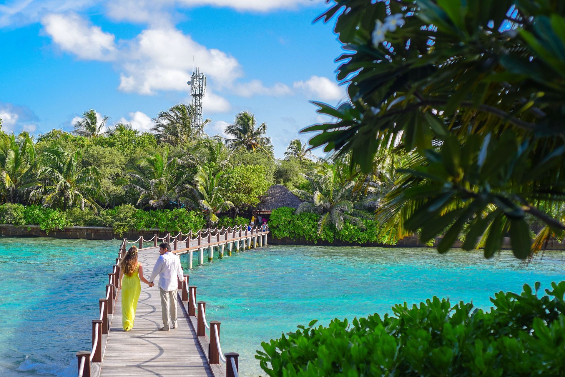 Couple on a wooden bridge over turquoise water in a lush tropical resort, under a bright blue sky