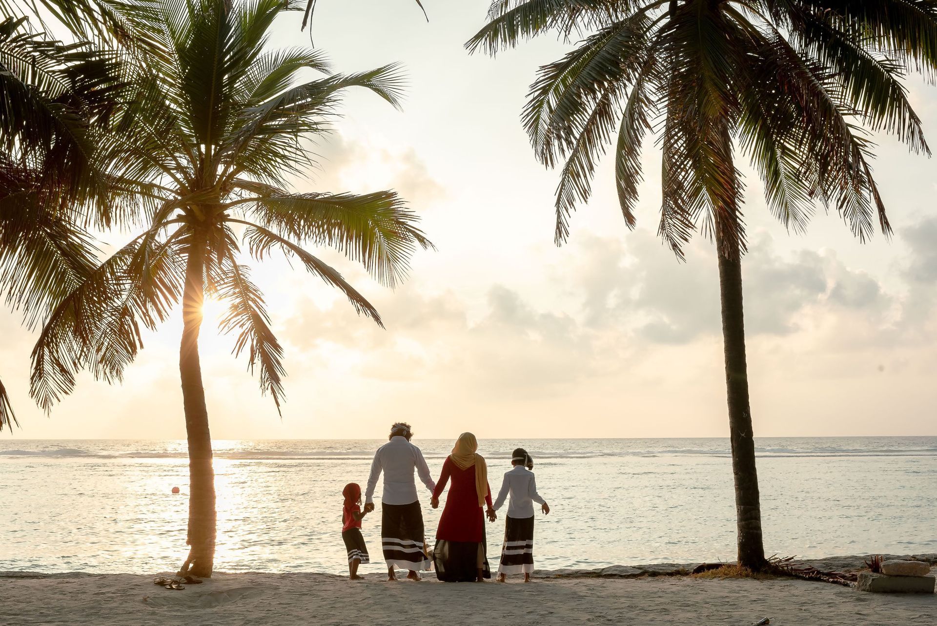 Family walking on a beach at sunset, framed by palm trees and calm ocean water.