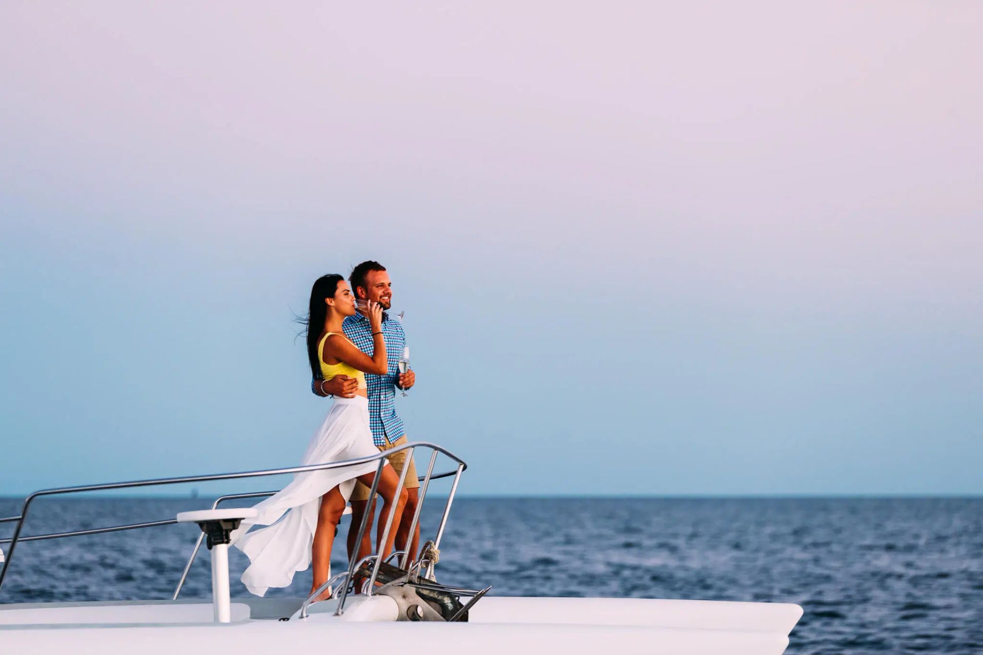 A couple stands at the bow of a boat on the open ocean, looking forward toward the horizon during a sunset.