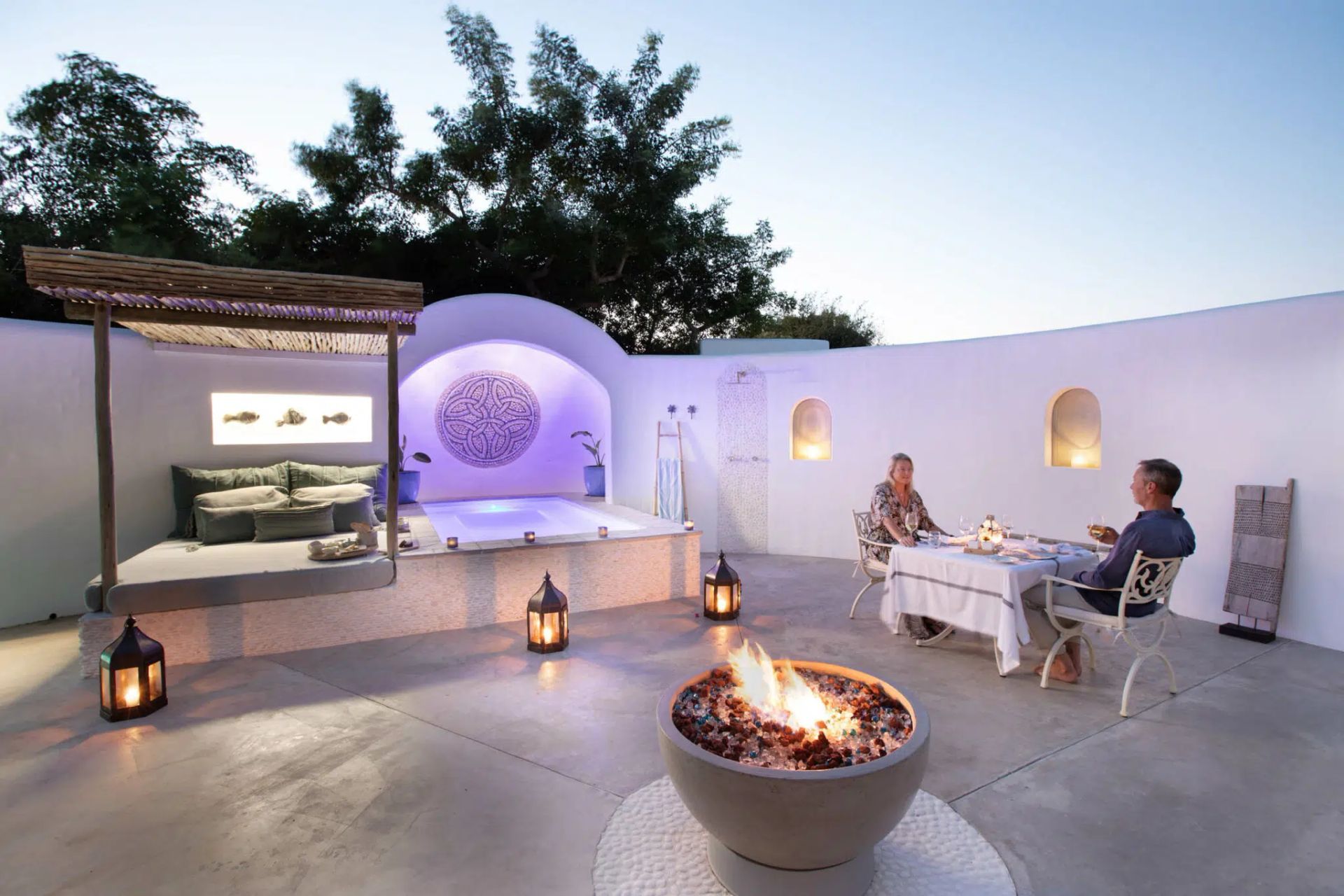 Outdoor patio with lanterns, fire pit, and two people seated at a table beside a white stucco wall