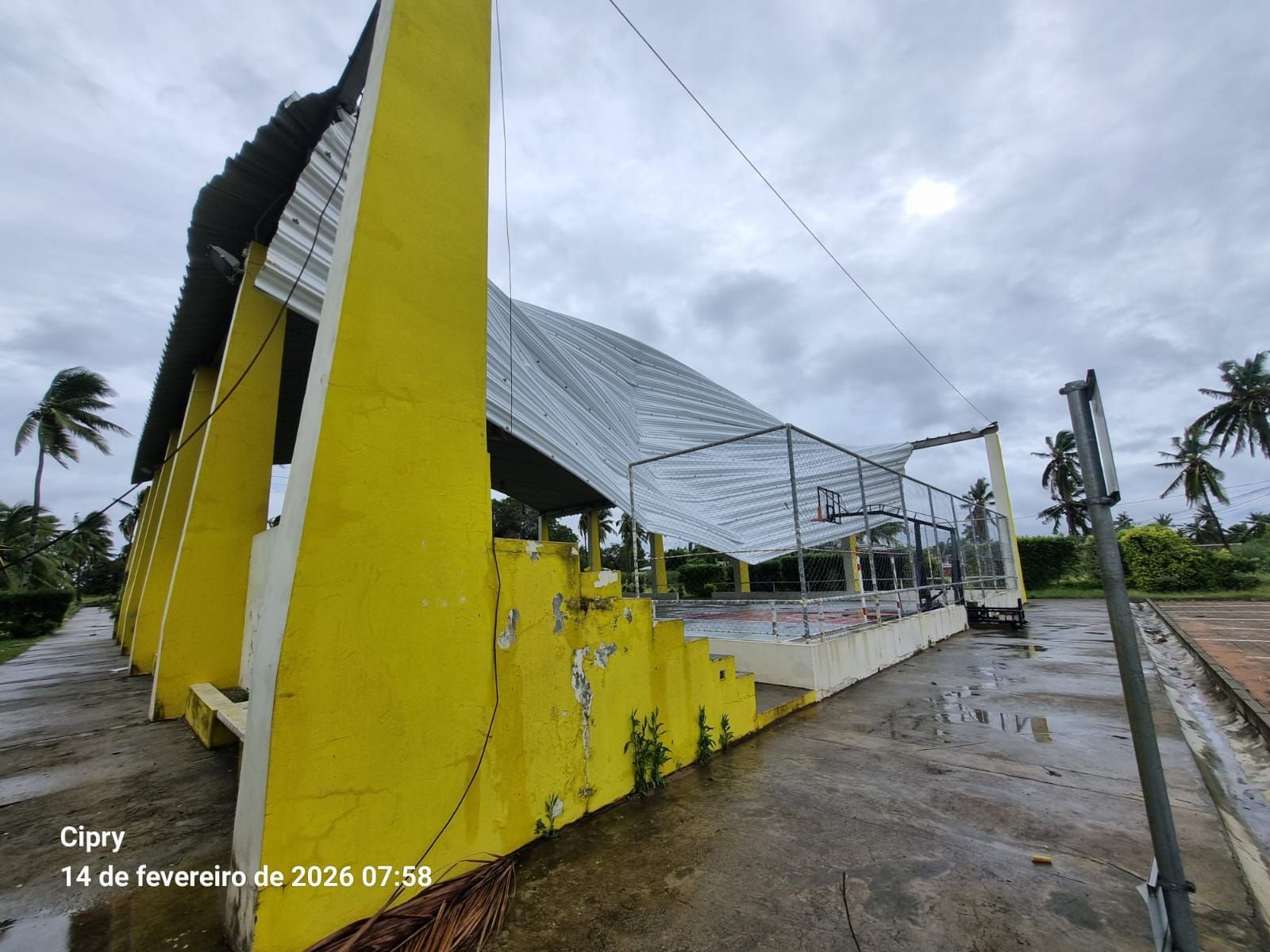 Yellow building with a metal roof on a wet roadside under a cloudy sky, with a walkway and palm trees nearby