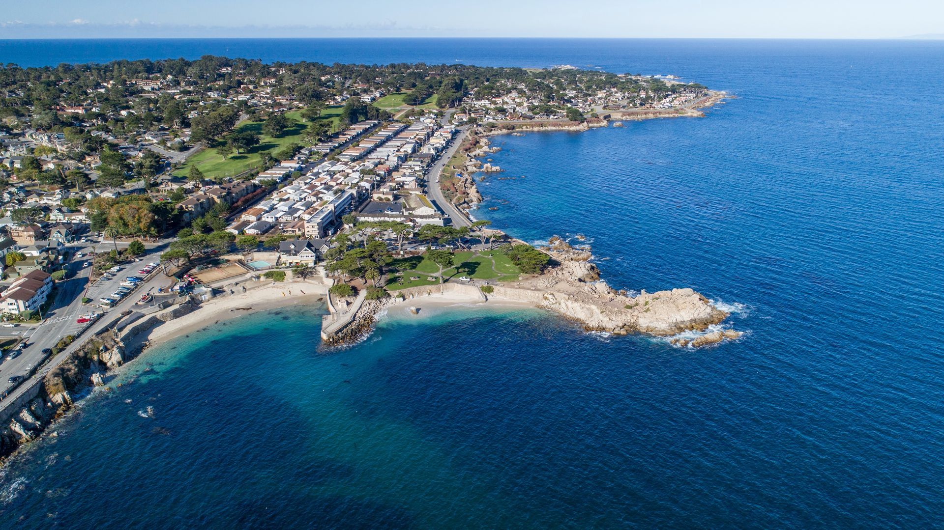 Aerial view of a coastal town featuring houses, a small sandy beach, and a rocky point stretching into the blue ocean.