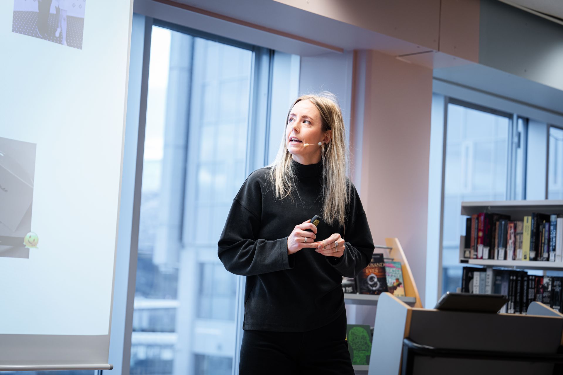Woman giving a presentation in a library. She is blonde and wearing a black sweater, gesturing with hands.