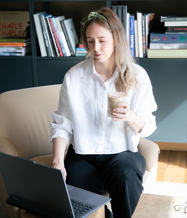 Dame sitter i en stol og holder et glass med kaffe. Hun ser på en laptop.