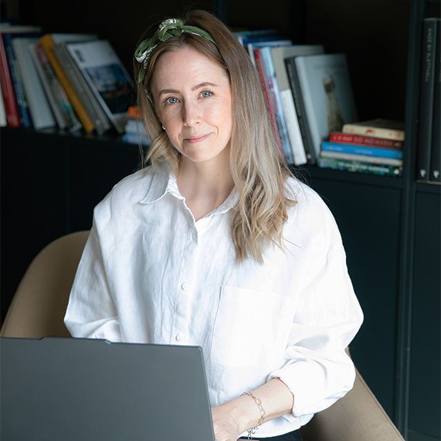 Monica Myklebust med hvit skjorte og laptop på fanget. Smiler til kamera.