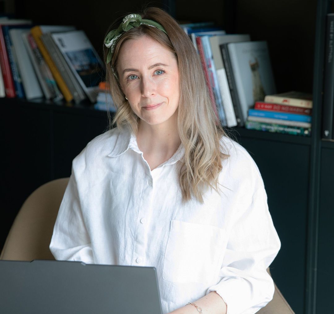 Monica Myklebust med hvit skjorte og laptop på fanget. Smiler til kamera.