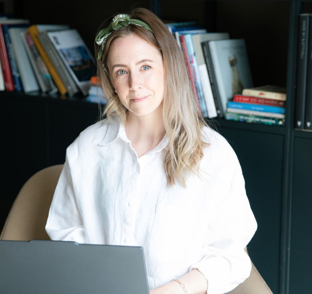 Monica Myklebust med hvit skjorte og laptop på fanget. Smiler til kamera.