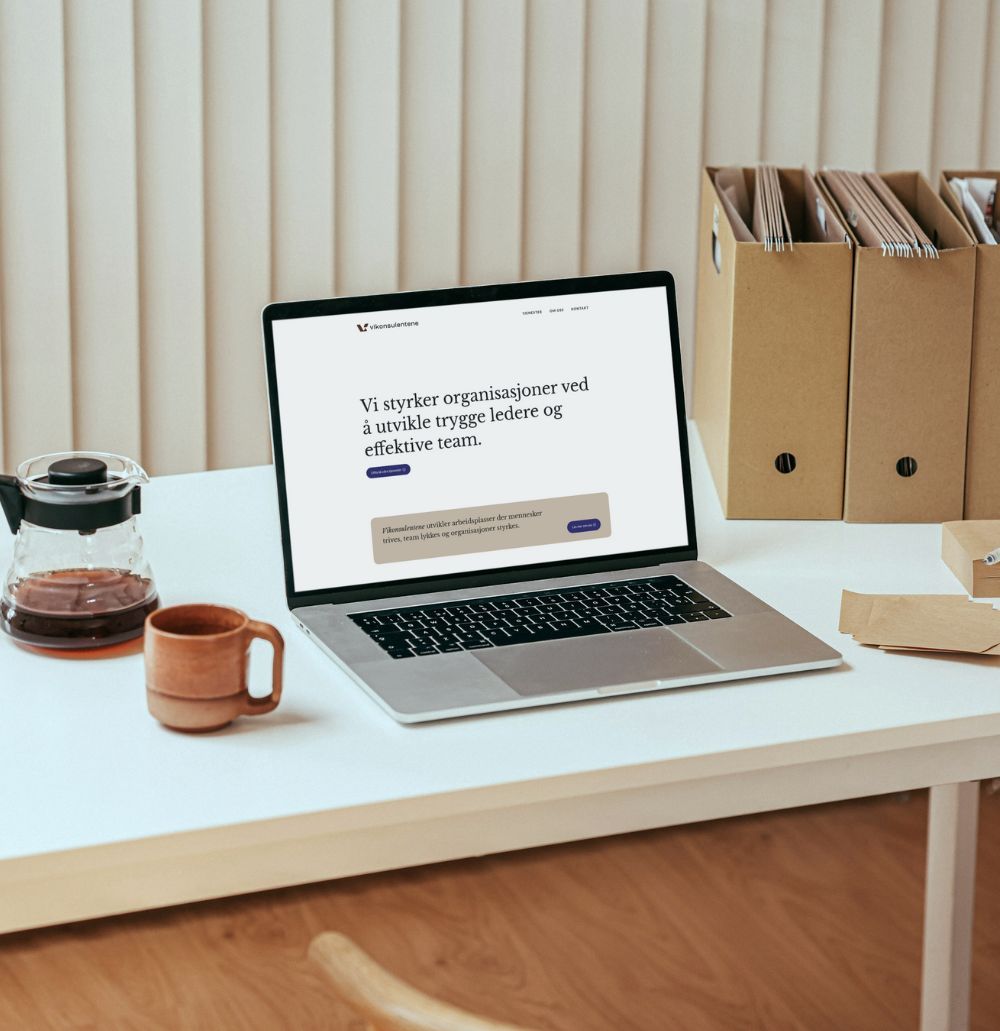 Laptop på kontorpult med nettside åpen. Kaffe  og permer på pulten.