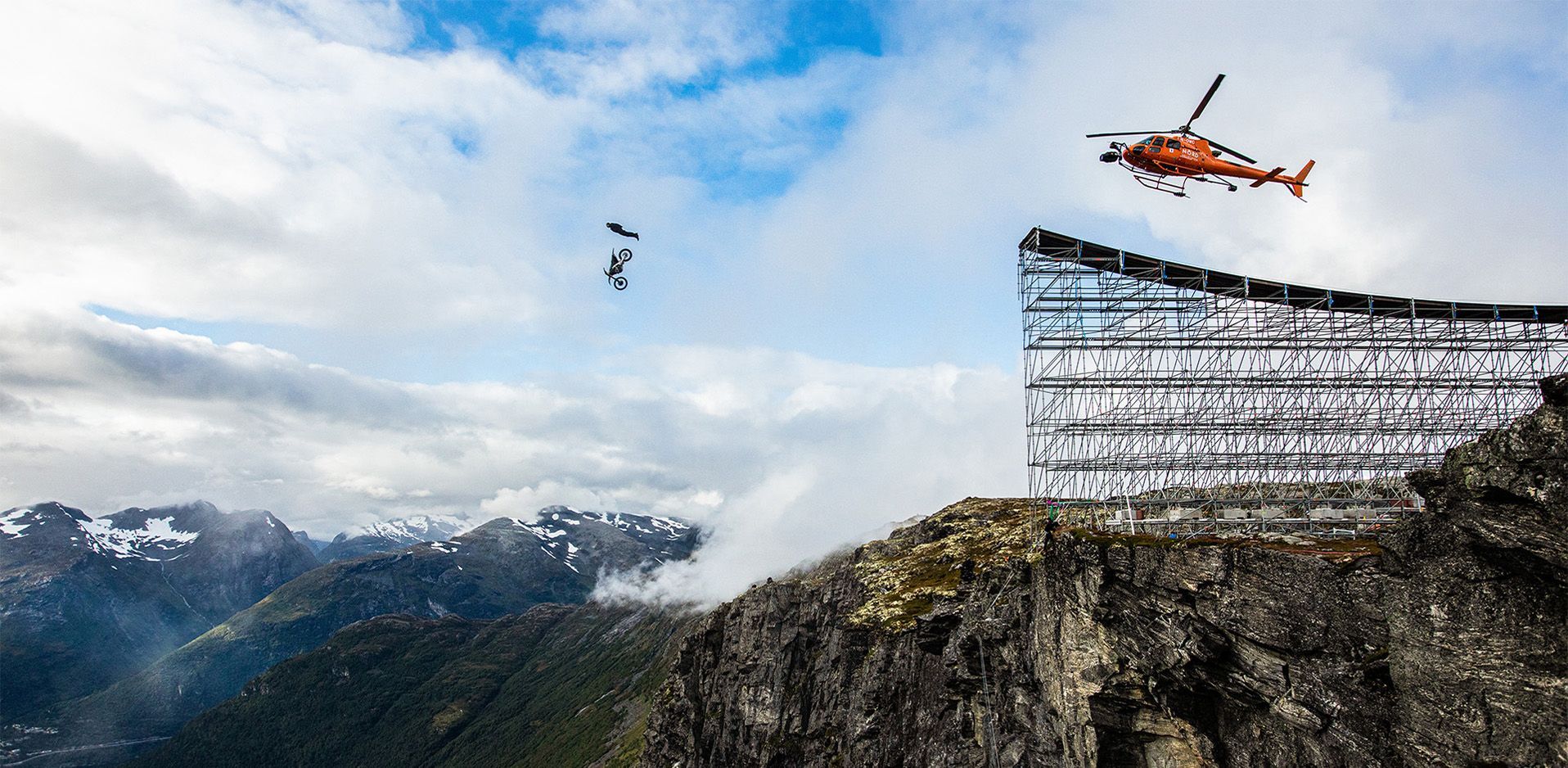 Tom Cruise on motorcycle in the air. Orange helicopter flying over.