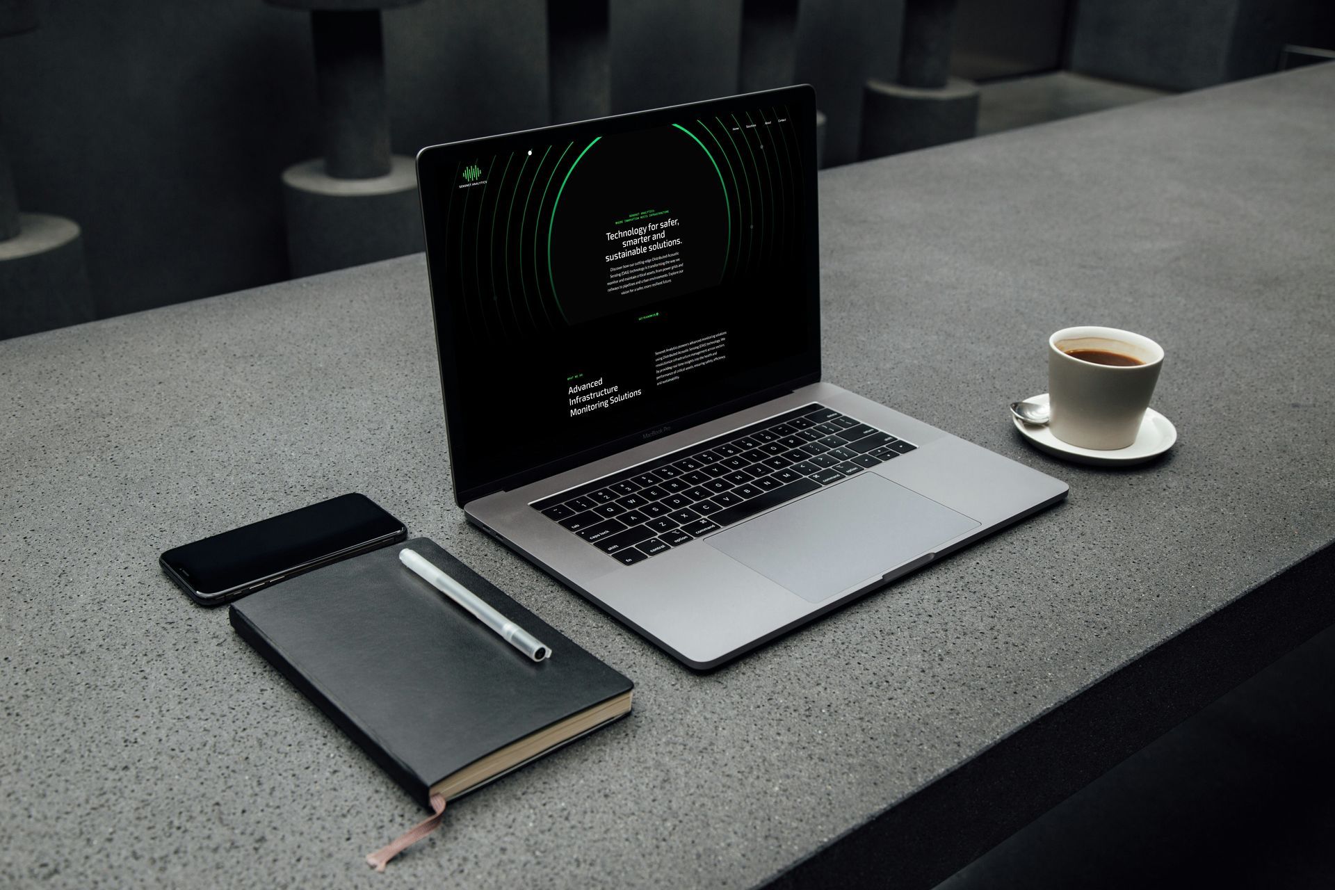 Laptop, kaffe, notatbok og telefon på mørkegrå kontorpult.