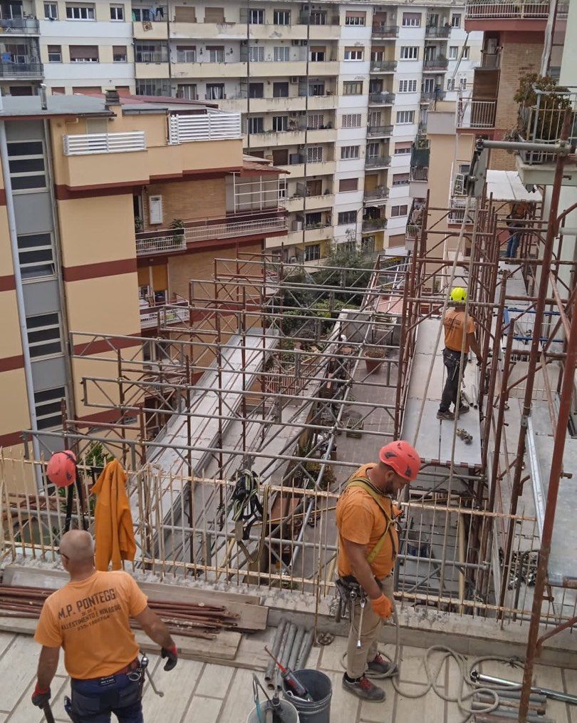 Operai edili su impalcature, impegnati nella riparazione della facciata di un edificio. Gli operai indossano dispositivi di sicurezza, magliette arancioni e caschi protettivi.