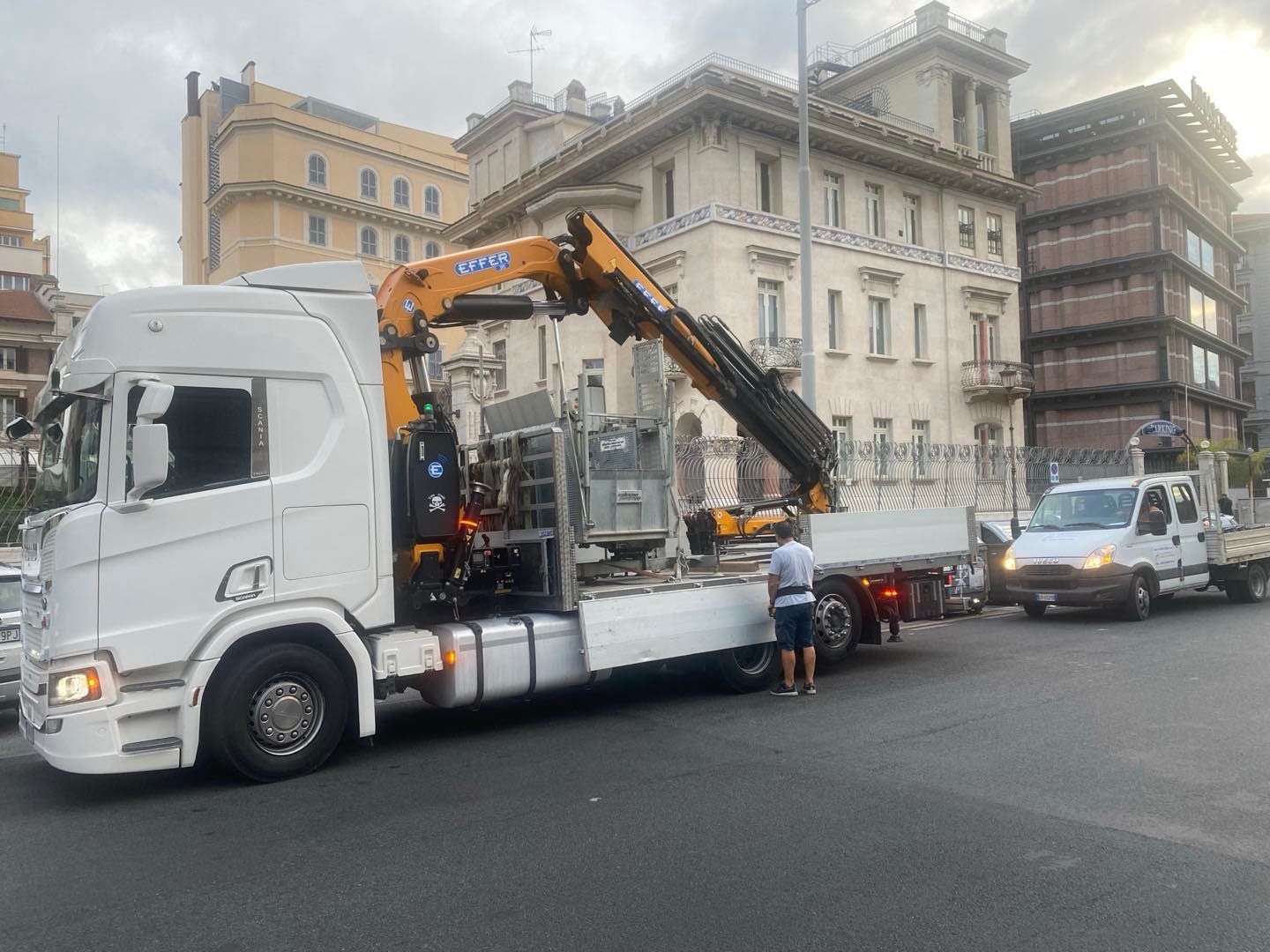Un camion bianco con una gru sta sollevando qualcosa su un pianale in una strada cittadina.