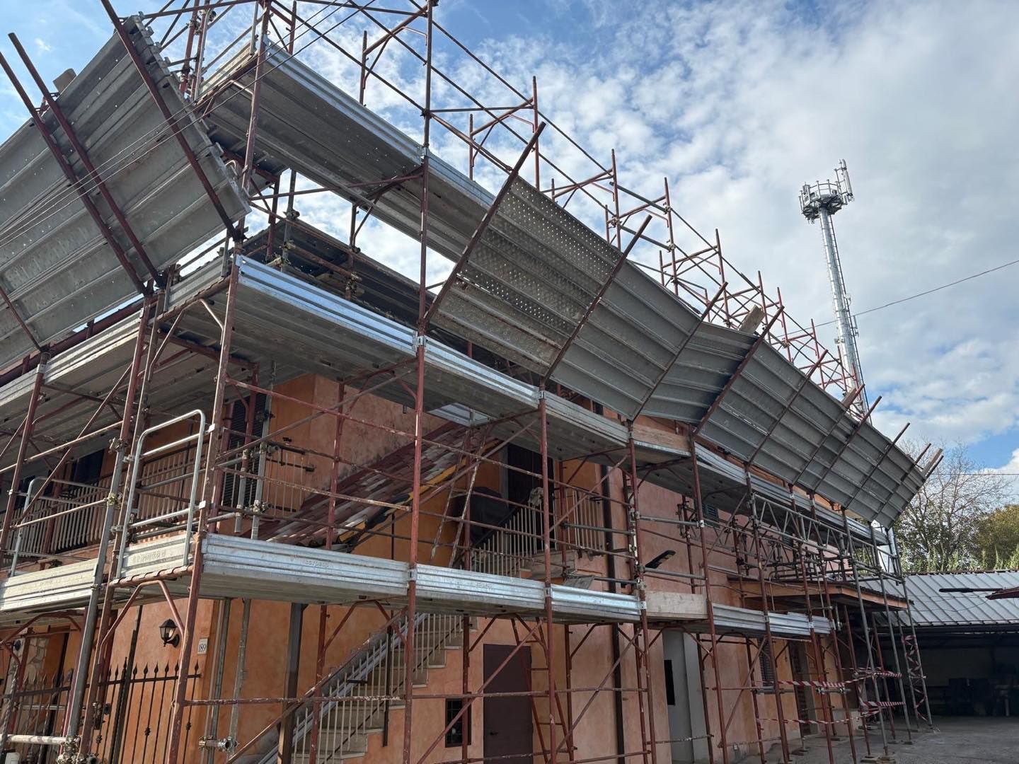 Un'impalcatura circonda un edificio a due piani. Lamiere metalliche ricoprono alcune parti della struttura, con un cielo nuvoloso sullo sfondo.