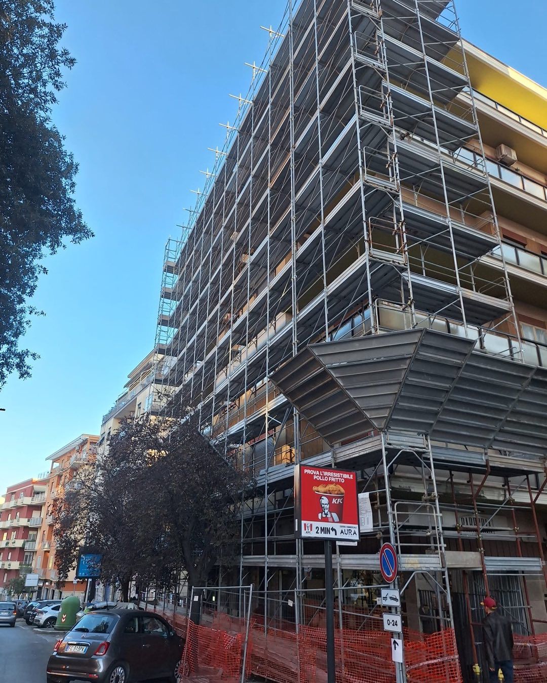 Edificio in fase di ristrutturazione con impalcature; vista sulla strada, recinzione di sicurezza rossa.