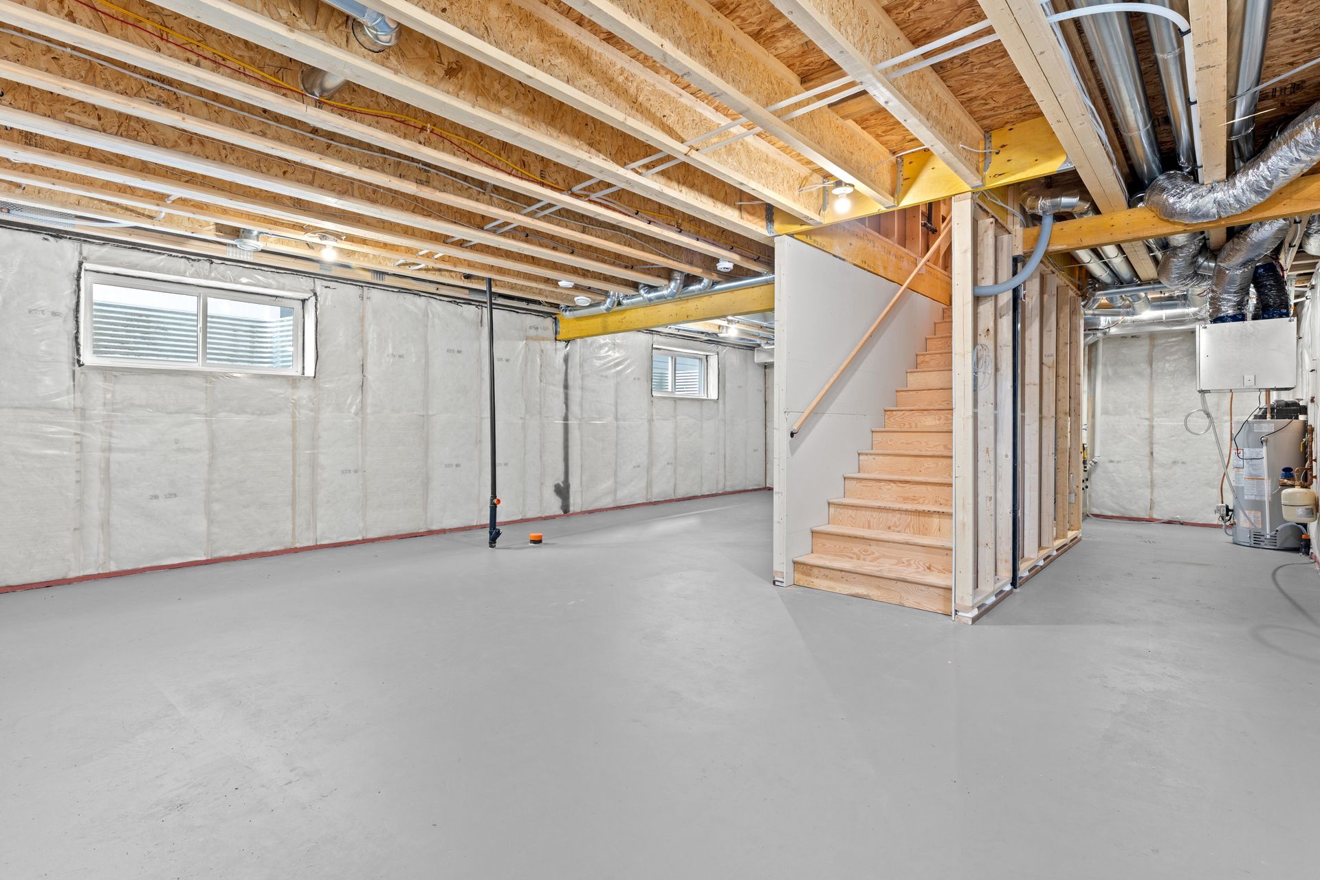 An unfinished basement with insulated walls, a grey concrete floor, exposed wooden ceiling beams, and a wooden staircase. An unfinished basement with insulated walls, a grey concrete floor, exposed wooden ceiling beams, and a wooden staircase.
