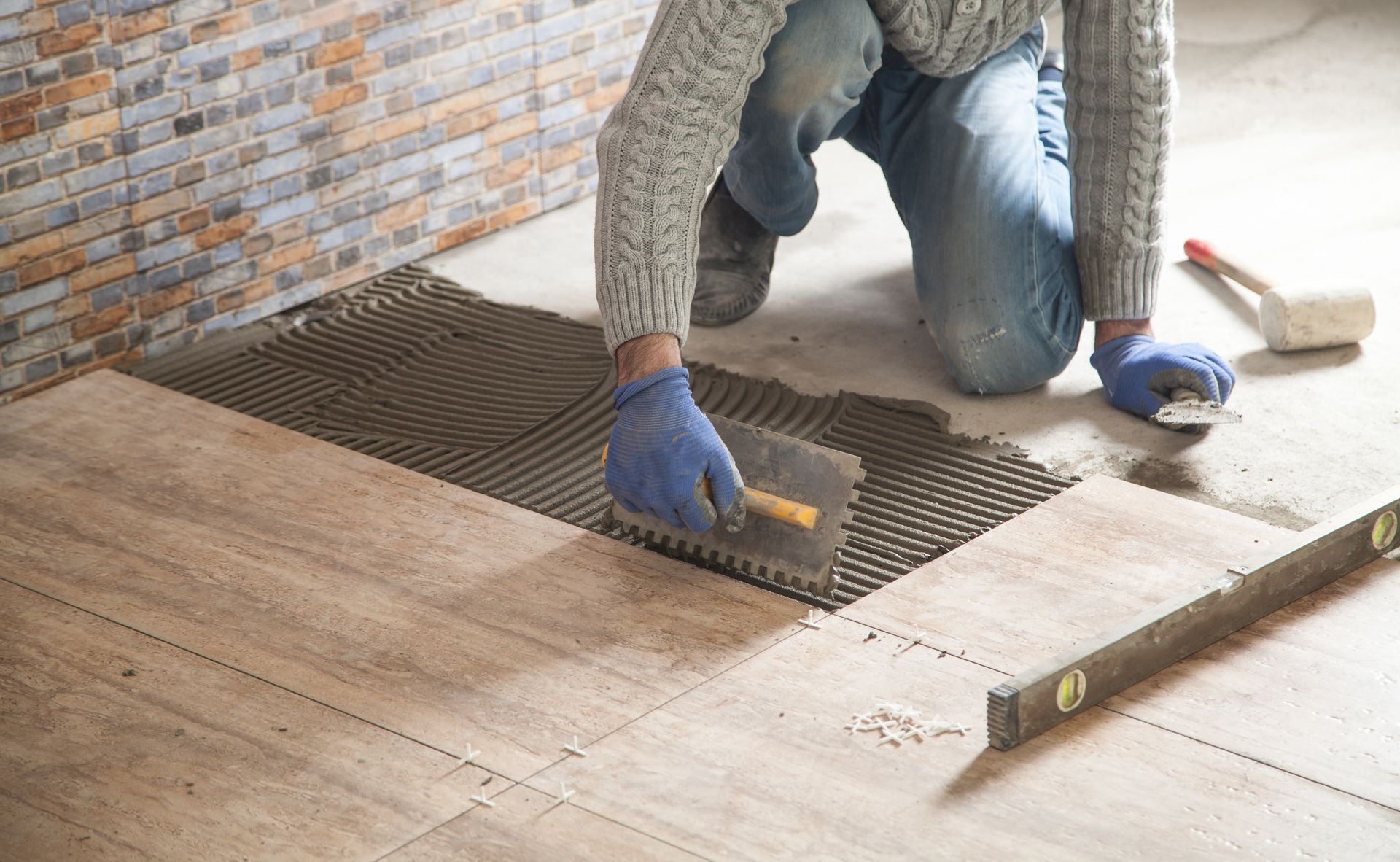 A person wearing blue gloves spreads mortar on a floor with a notched trowel, preparing to install wood-look tile. A person wearing blue gloves spreads mortar on a floor with a notched trowel, preparing to install wood-look tile.