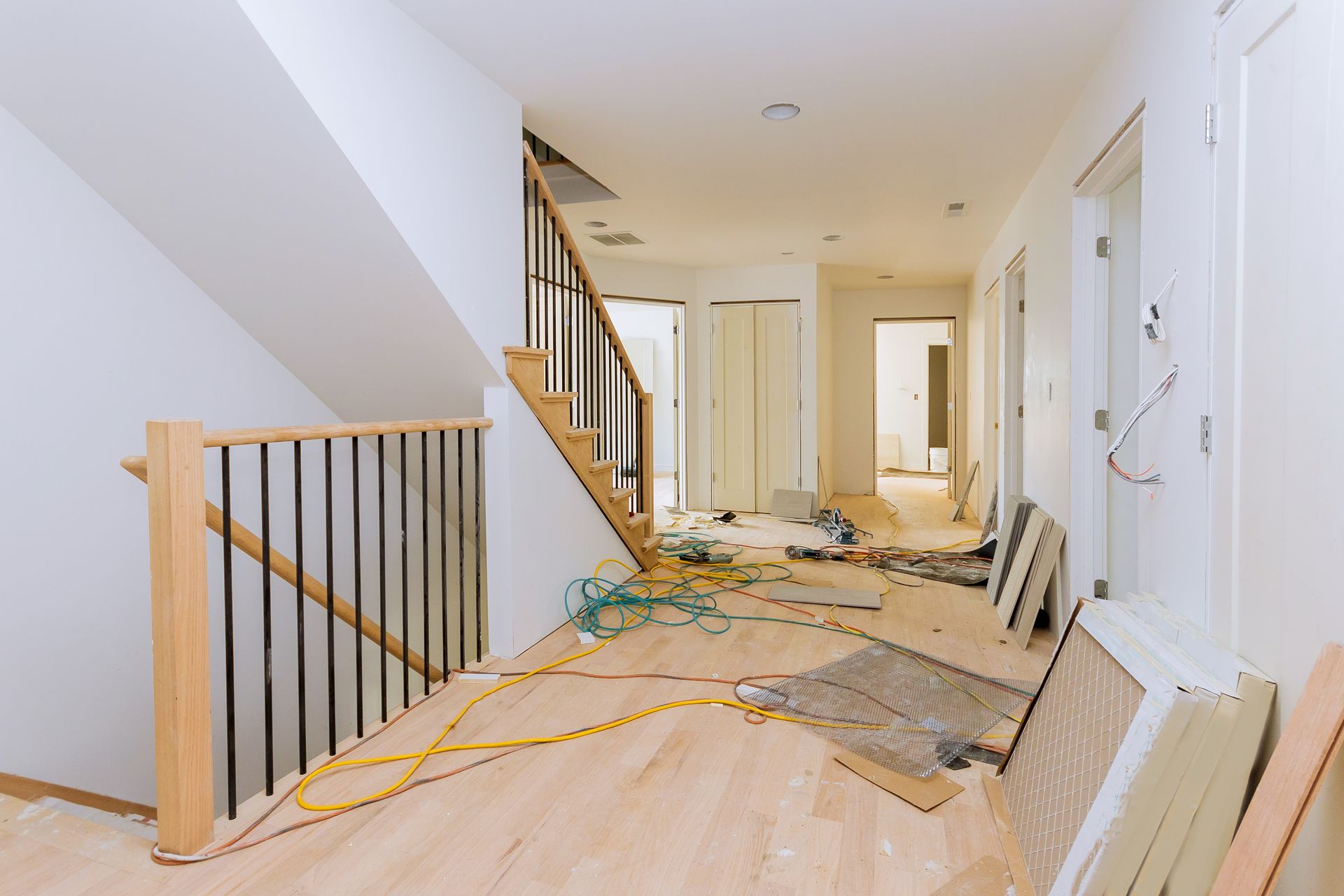 A hallway in a house under renovation with light wood flooring, an open staircase, exposed wiring, and construction debris. A hallway in a house under renovation with light wood flooring, an open staircase, exposed wiring, and construction debris.